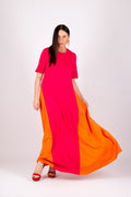 A woman modeling a long, flowing color-block dress with bright pink on the upper portion and orange on the lower portion, paired with red heels, against a plain light gray studio background.
