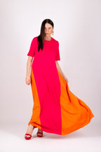 A woman modeling a long, flowing color-block dress with bright pink on the upper portion and orange on the lower portion, paired with red heels, against a plain light gray studio background.
