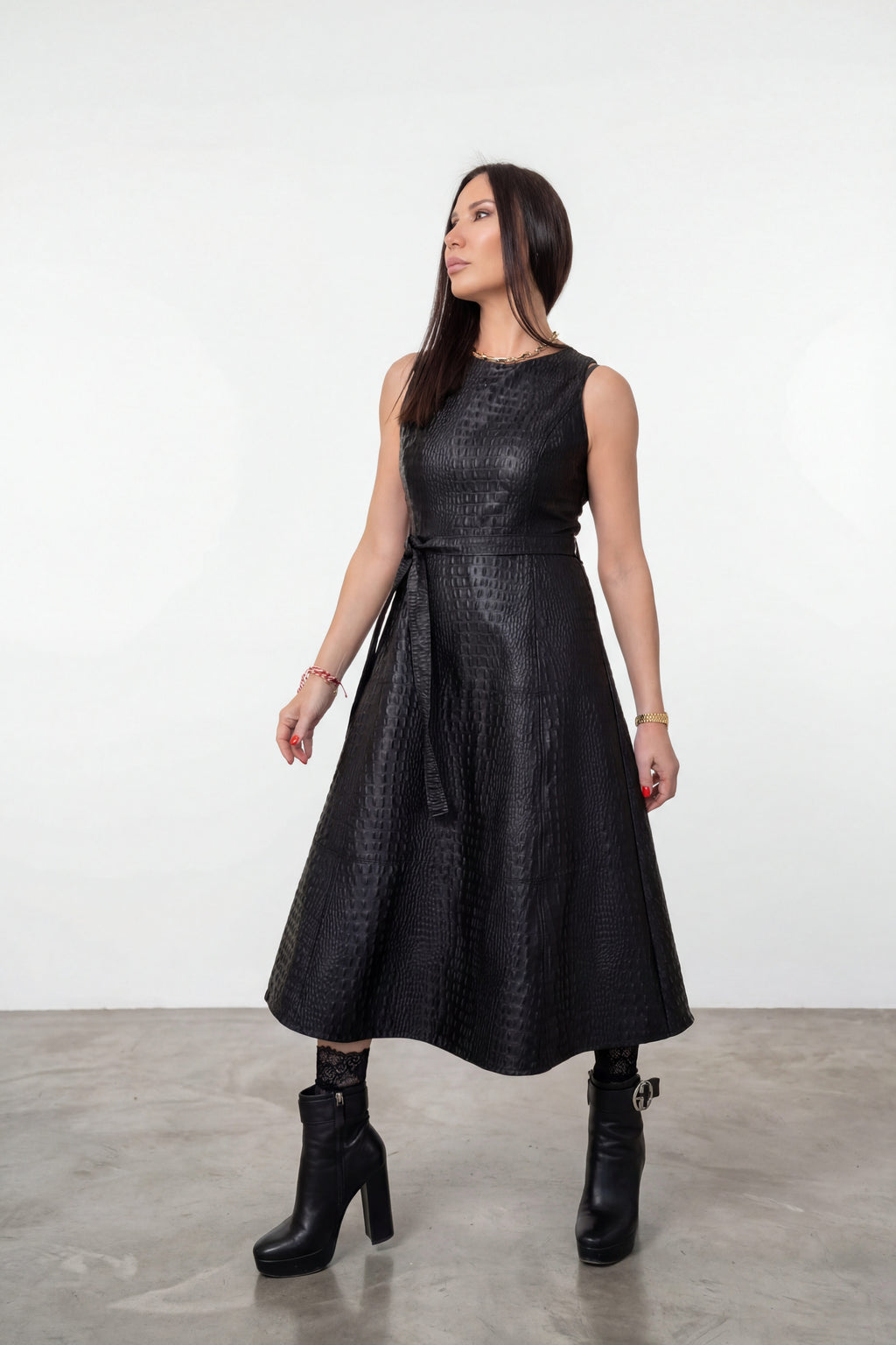 A woman modeling a sleeveless black textured faux-leather midi dress with a tie waist, paired with black ankle boots, in a studio setting with a white wall and gray floor.