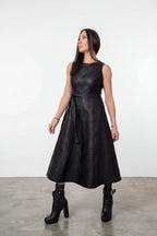 A woman modeling a sleeveless black textured faux-leather midi dress with a tie waist, paired with black ankle boots, in a studio setting with a white wall and gray floor.