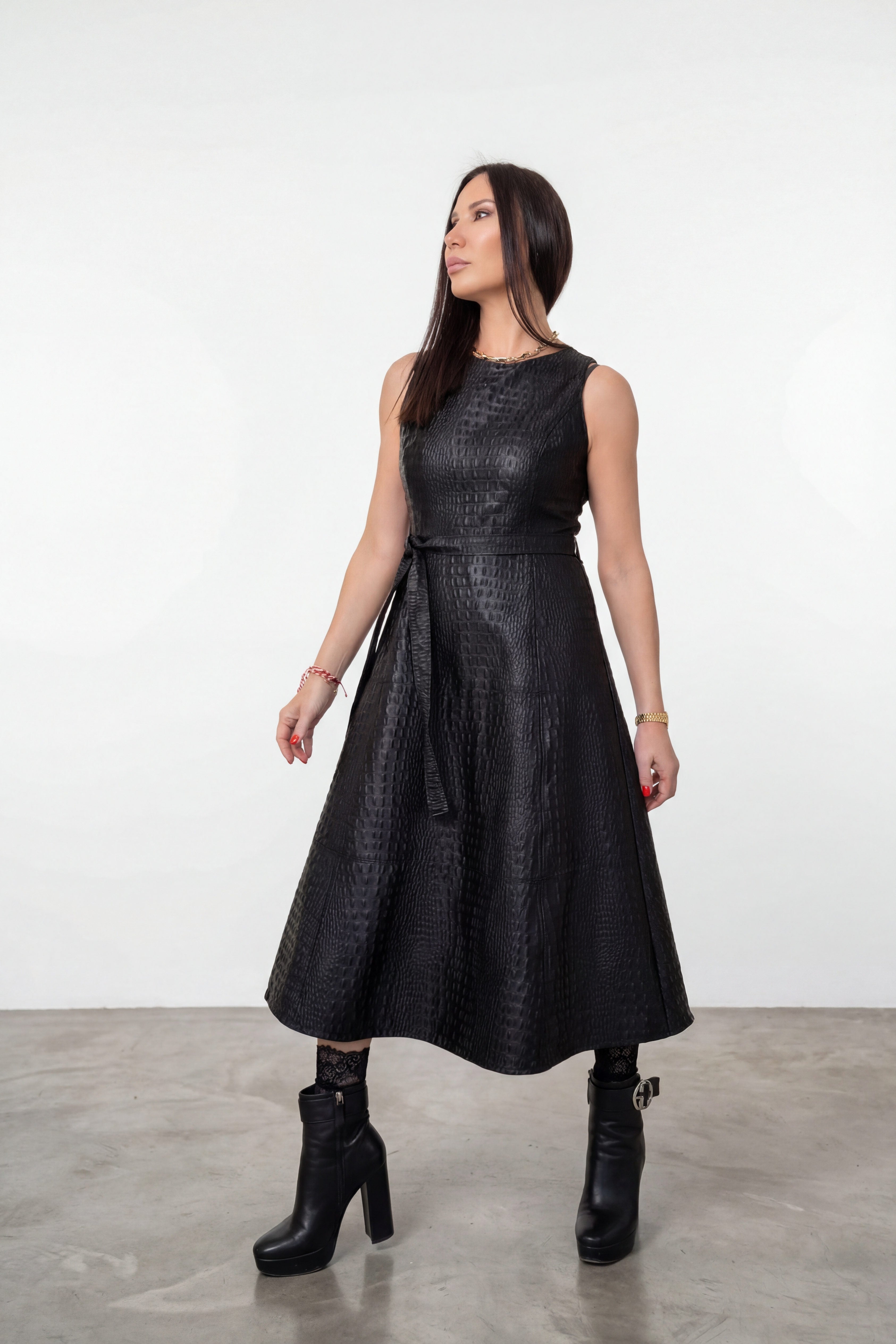 A woman modeling a sleeveless black textured faux-leather midi dress with a tie waist, paired with black ankle boots, in a studio setting with a white wall and gray floor.
