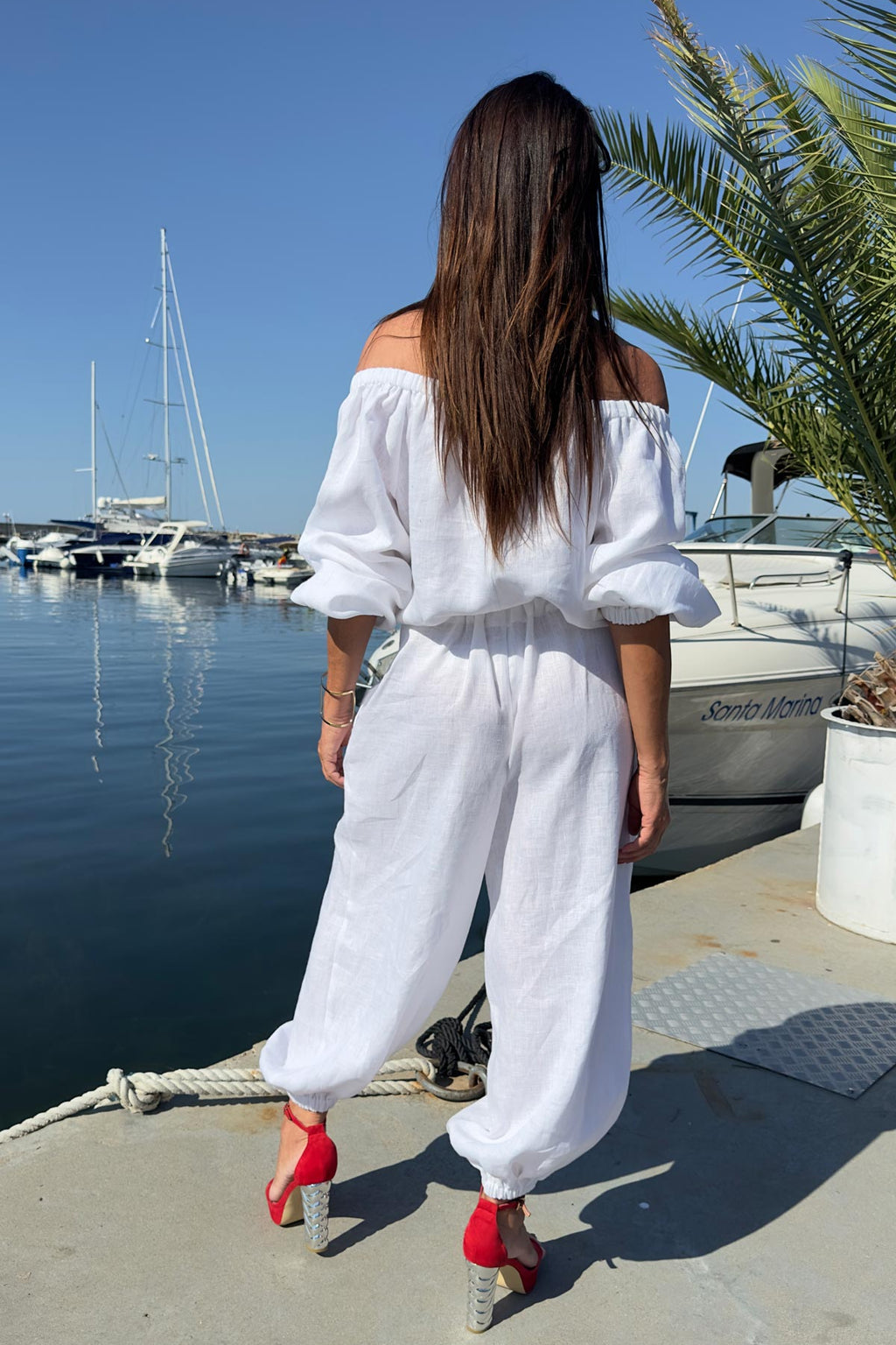 Woman in a white outfit standing by a marina with boats and palm trees.