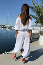 Woman in a white outfit standing by a marina with boats and palm trees.