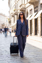 A woman walking on a cobblestone city street wearing a navy blue pinstripe blazer and matching trousers, pulling a black rolling suitcase.