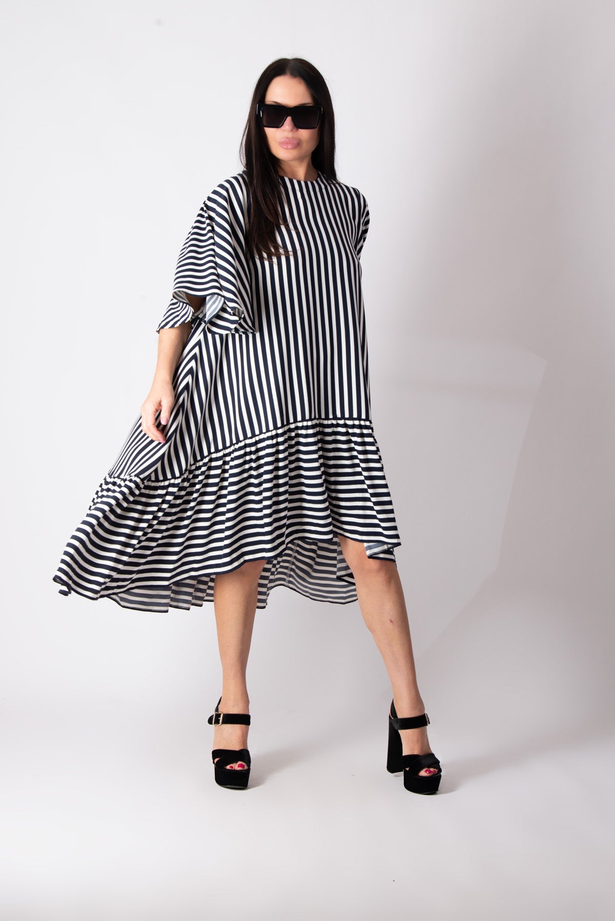 A woman modeling a black and white vertically striped midi dress with a ruffled hem and elbow-length sleeves, paired with black platform sandals against a plain light gray studio background.