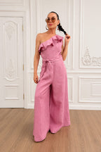 Woman wearing a pink one-shoulder jumpsuit in a room with white walls and wooden floor.