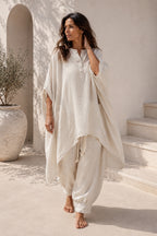 Woman in a flowing white outfit standing outdoors against a light-colored wall.