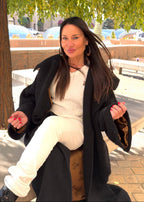 Woman sitting outdoors under a tree, wearing a black coat and white pants.