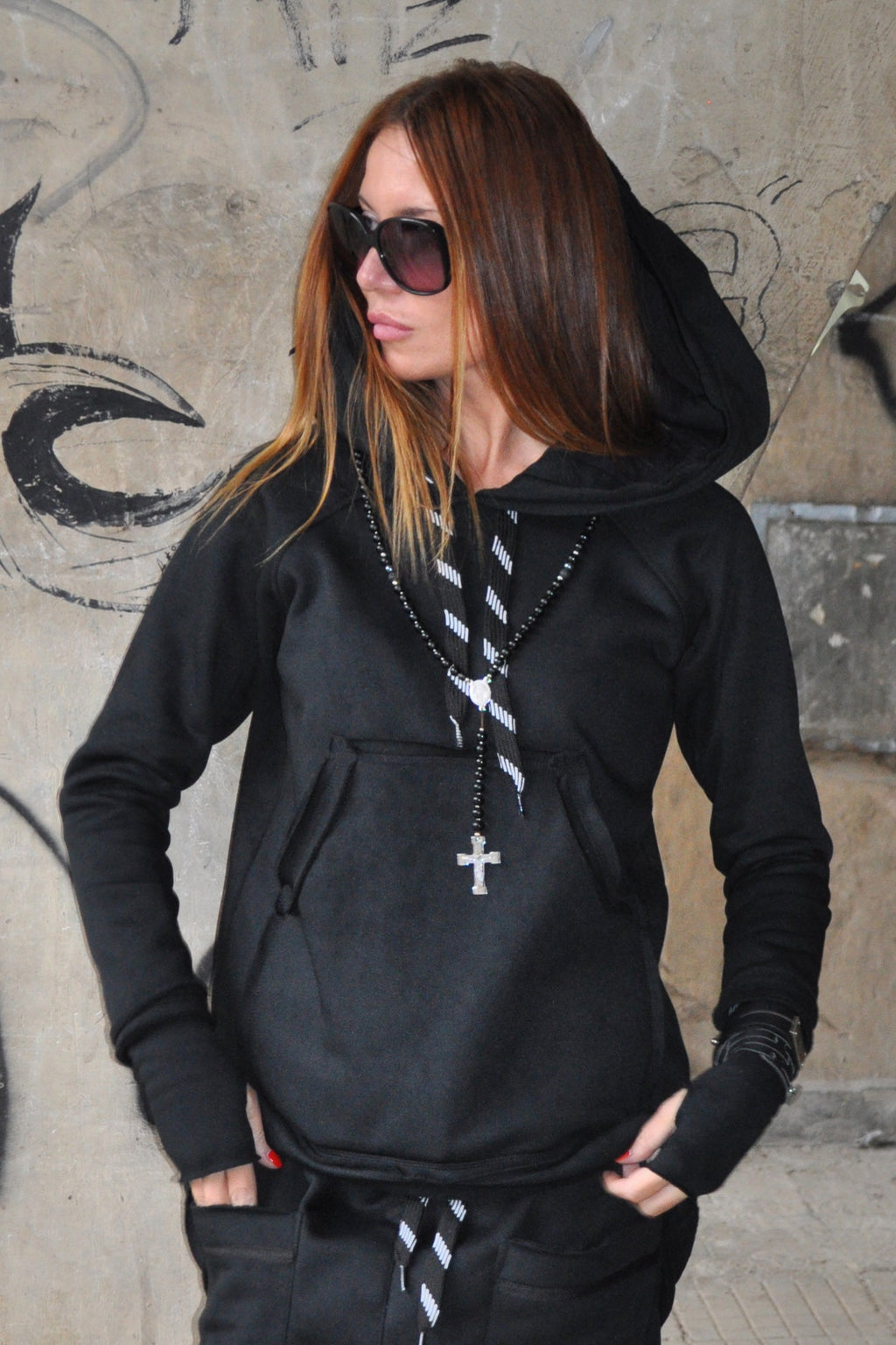 Person wearing a black hoodie with a cross necklace against a graffiti-covered wall.