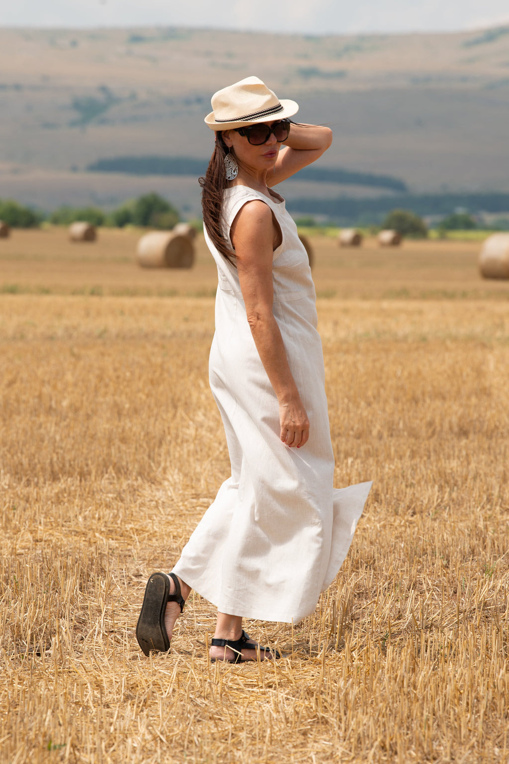 Woman in a white linen 
dress and hat standing in a field with hay bales.
