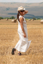 Woman in a white linen 
dress and hat standing in a field with hay bales.