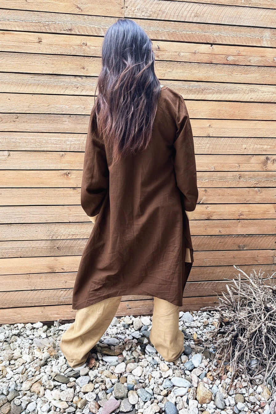 Person wearing a brown coat standing against a wooden wall with pebbles and plants on the ground.