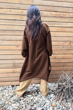 Person wearing a brown coat standing against a wooden wall with pebbles and plants on the ground.