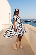 A woman wearing a black and white polka dot dress, beige wedge sandals, and sunglasses stands on a sunny balcony overlooking a blue sea with yachts.