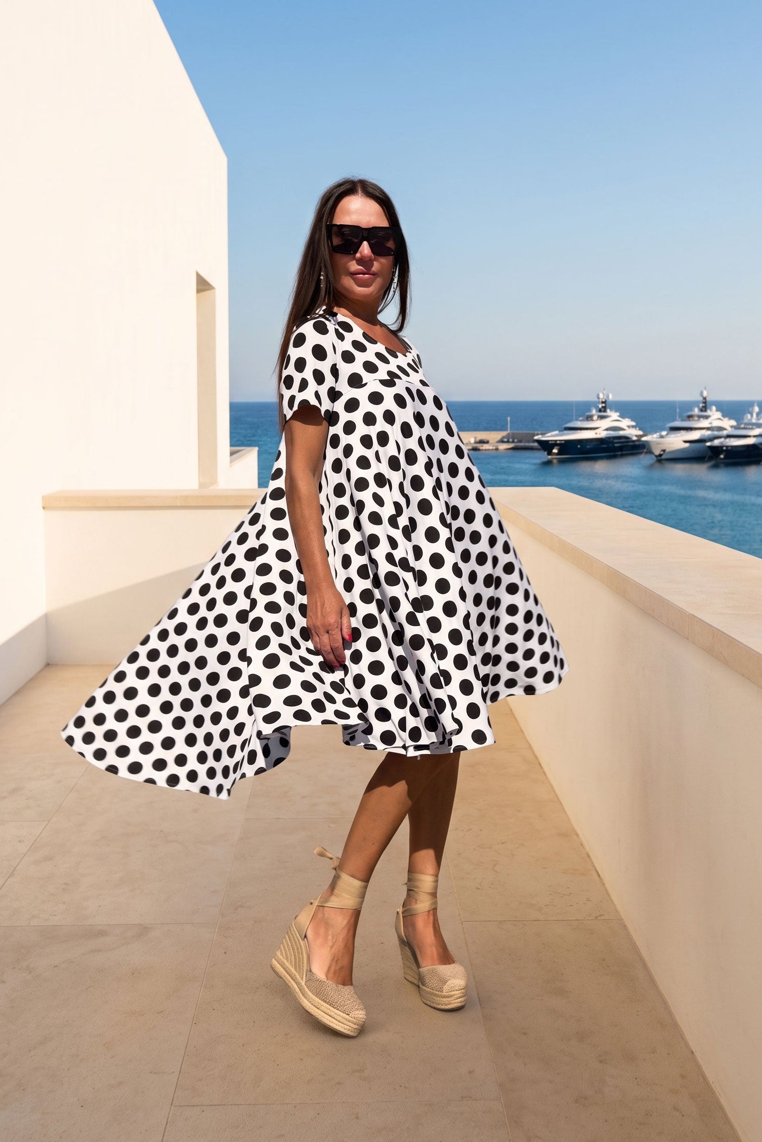 A woman wearing a black and white polka dot dress, beige wedge sandals, and sunglasses stands on a sunny balcony overlooking a blue sea with yachts.