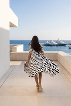 A woman wearing a black and white polka dot dress walks away on a sunlit terrace overlooking a blue sea with yachts in the distance.