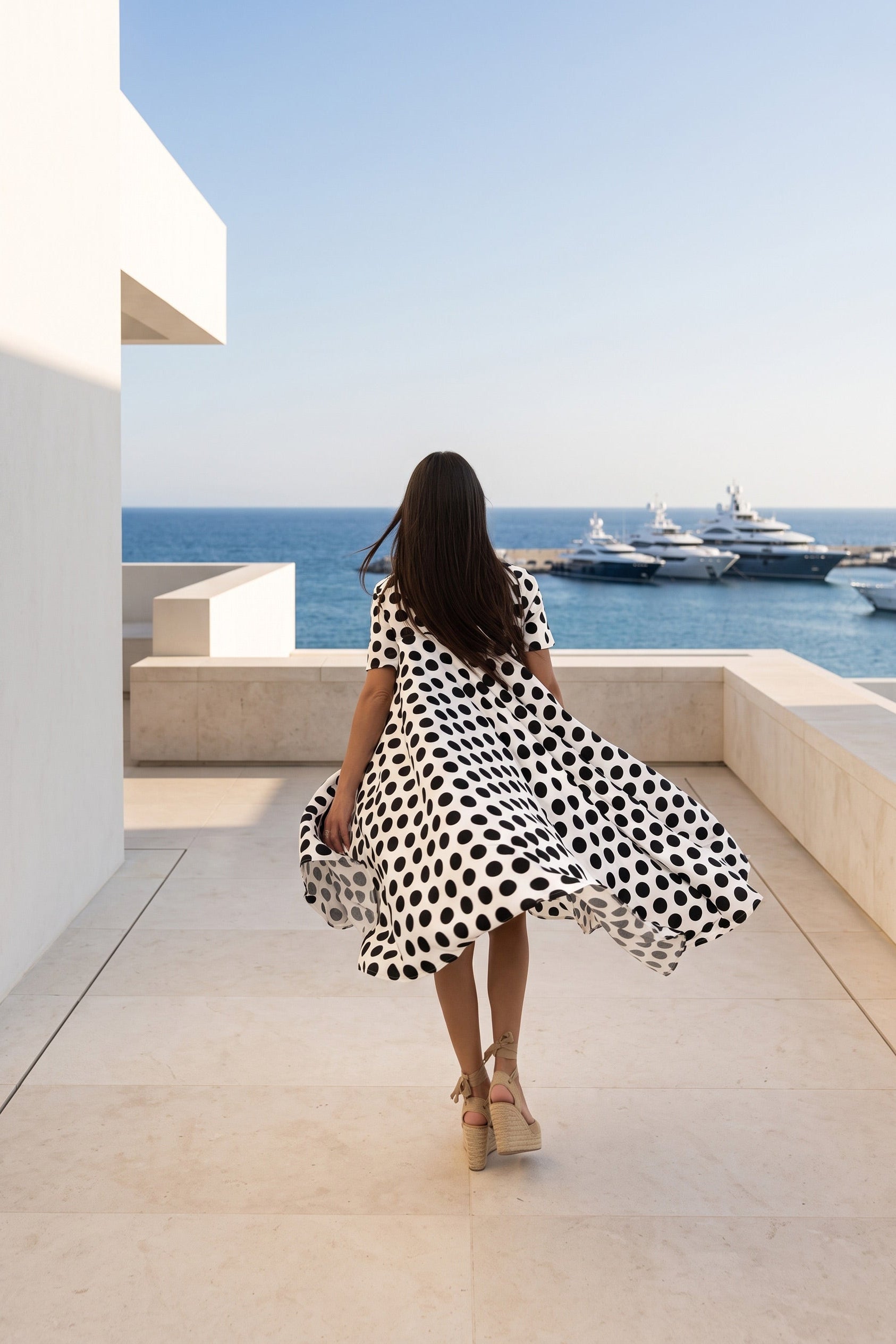A woman wearing a black and white polka dot dress walks away on a sunlit terrace overlooking a blue sea with yachts in the distance.