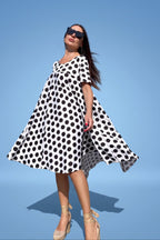 A woman wearing a black and white polka dot dress, black sunglasses, and beige high-heeled sandals stands against a solid blue background, looking upward with her dress spread out.