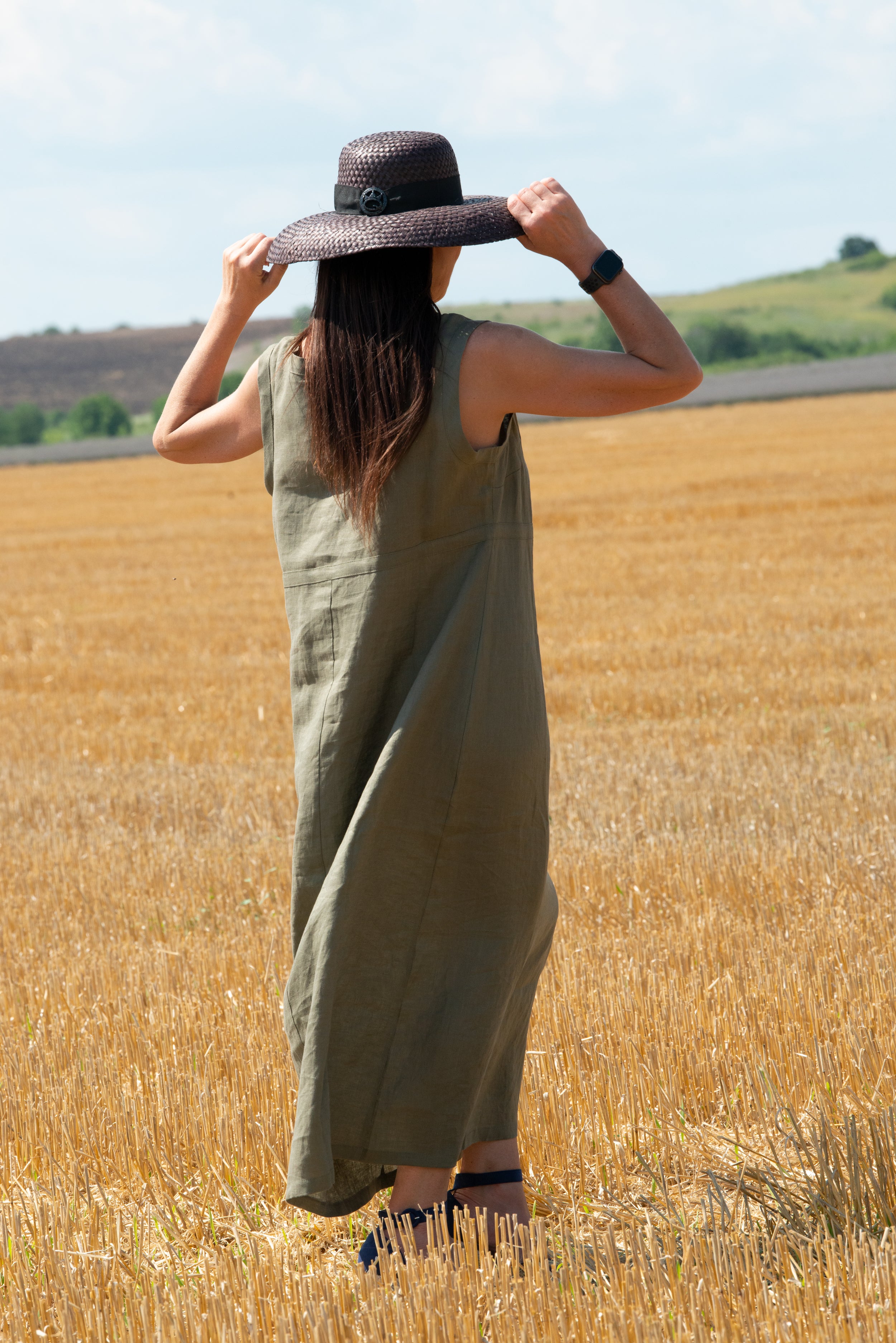 Woman in a long linen 
green dress and wide-brimmed hat standing in a wheat field.