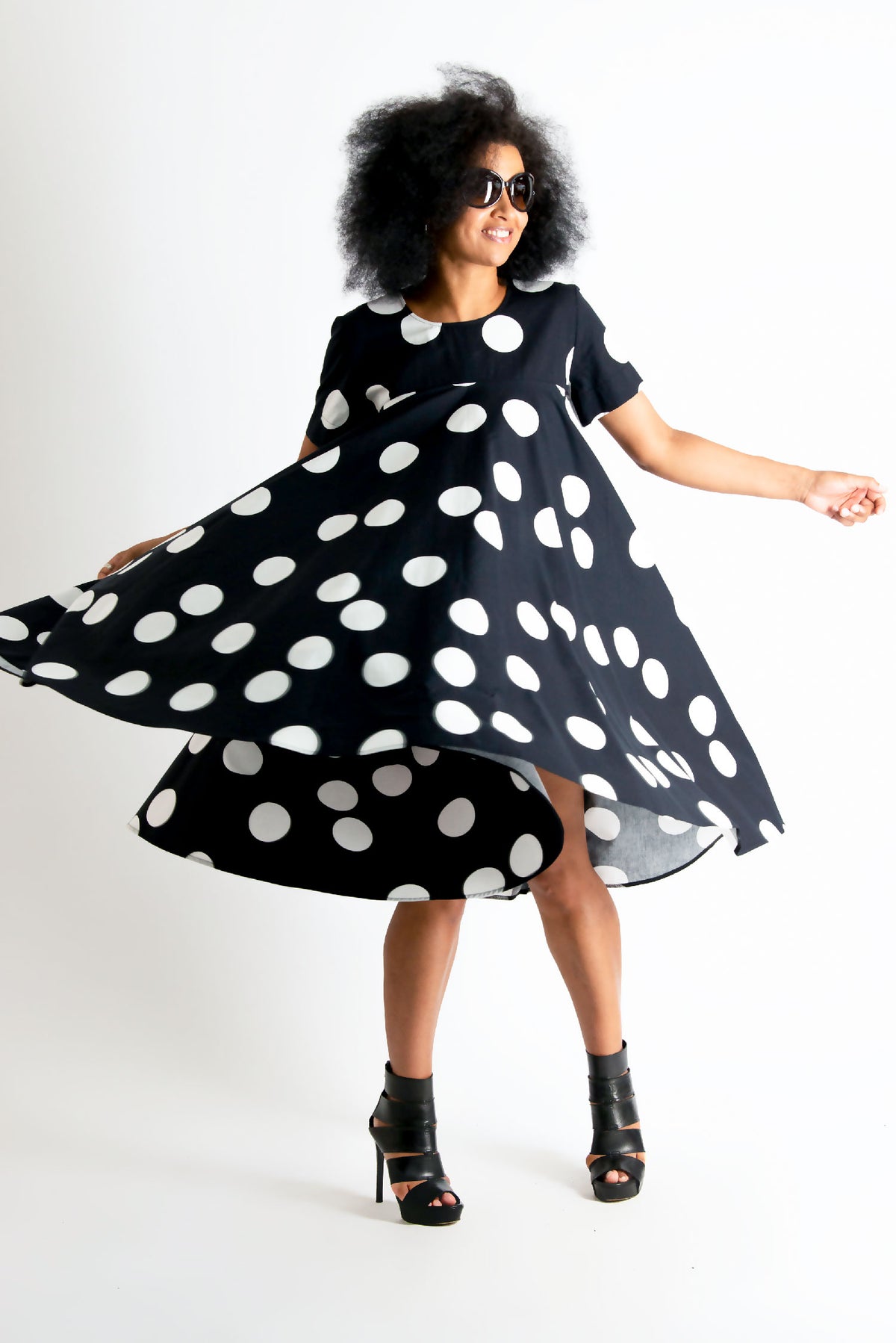 A woman wearing a black short-sleeve dress with large white polka dots, captured mid-spin to show the flared skirt. She is wearing black high-heeled sandals and sunglasses against a plain white studio background.