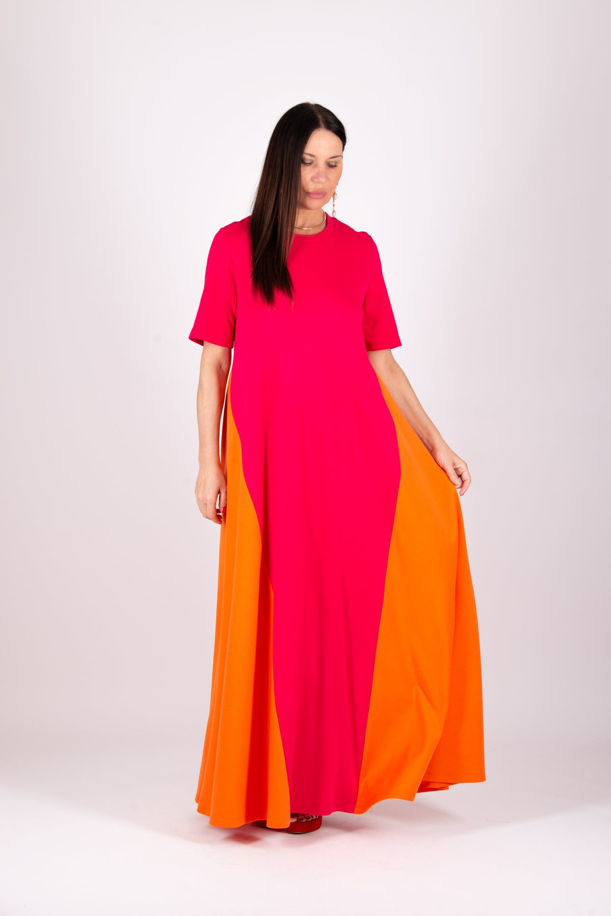 A woman modeling a vibrant color-blocked maxi dress with a bright pink upper section and a flowing orange skirt, set against a plain light gray studio background.