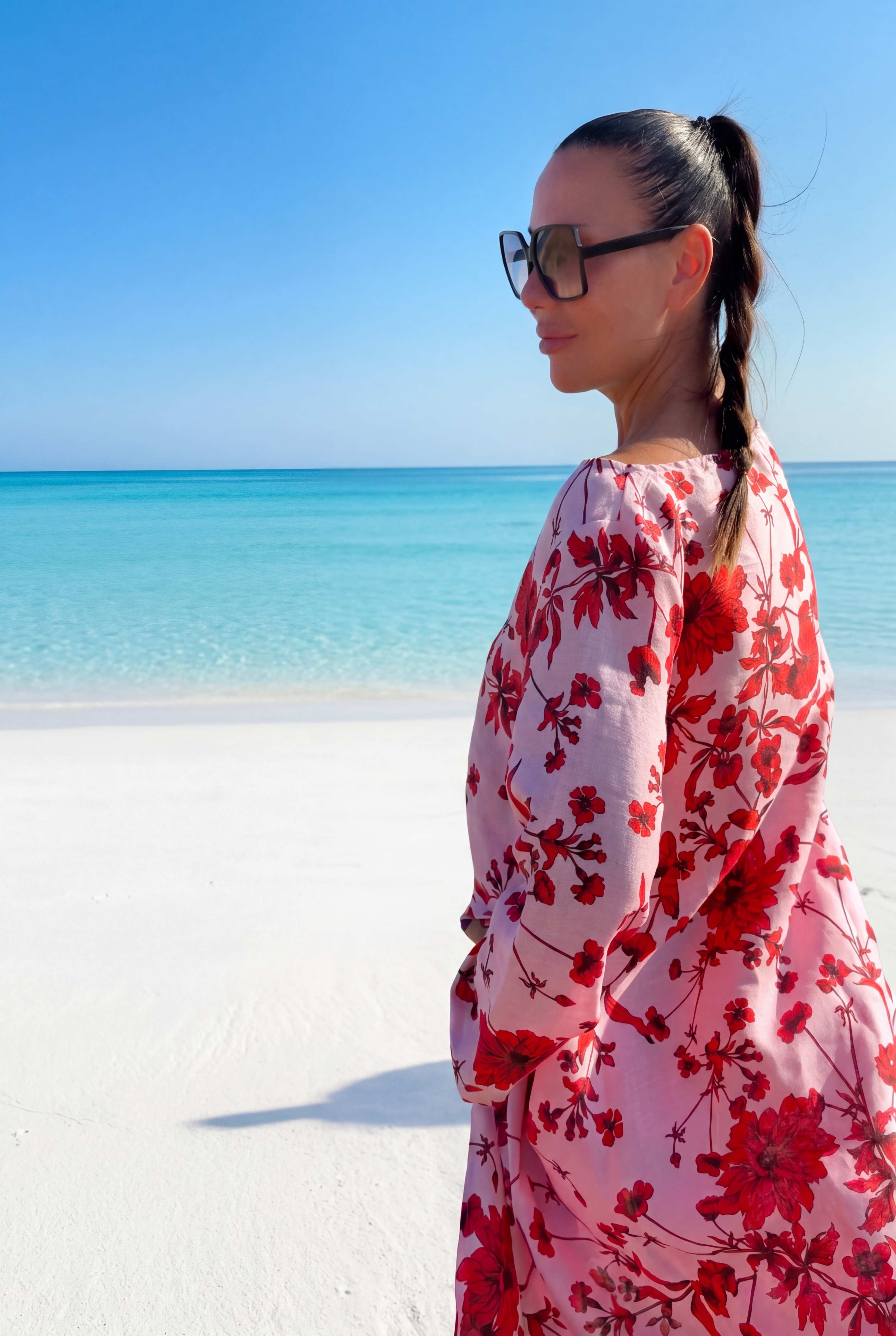 Woman in a red floral dress standing on a beach with clear blue water and sky.
