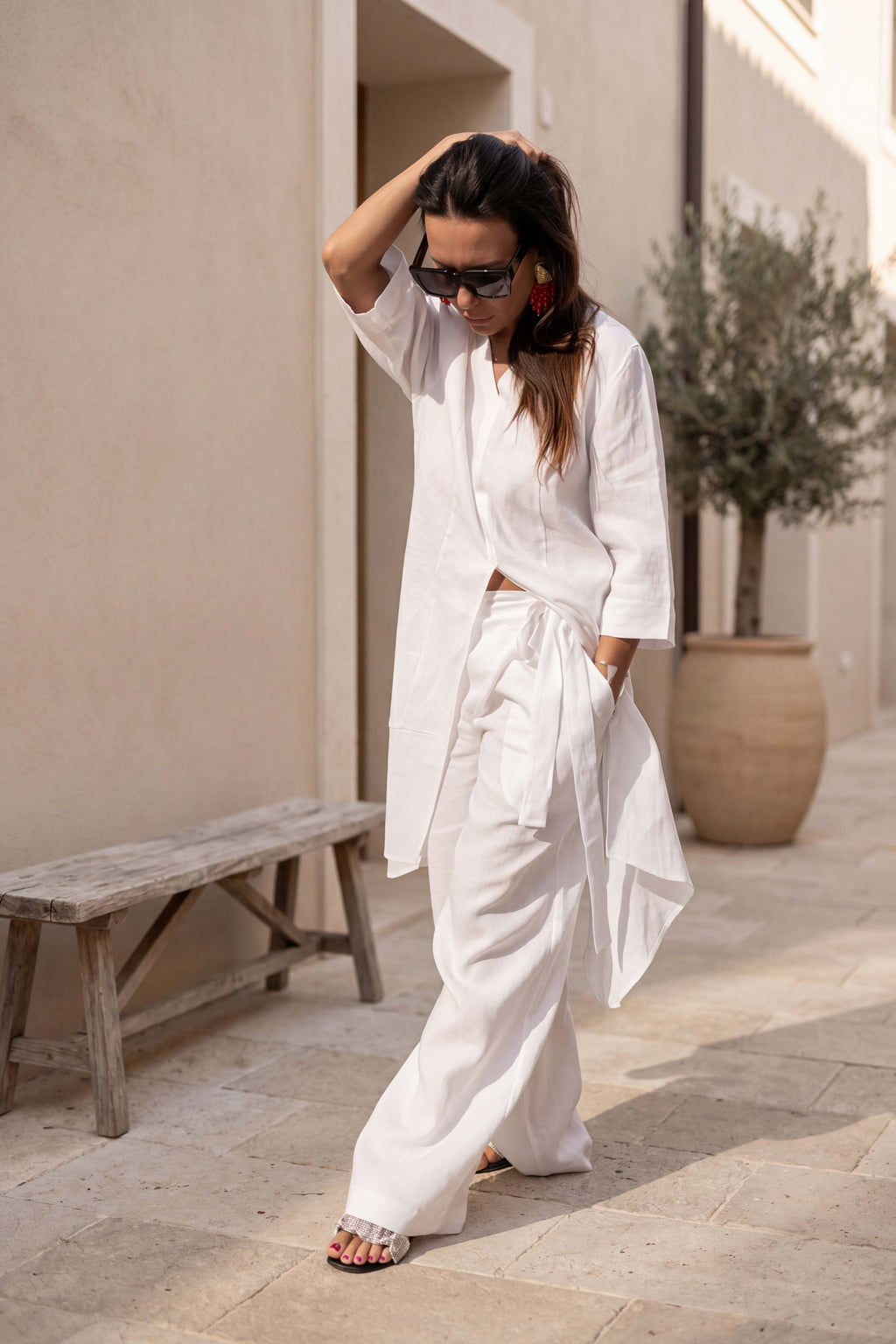 Woman in a white outfit standing outdoors near a building with a bench and plant.
