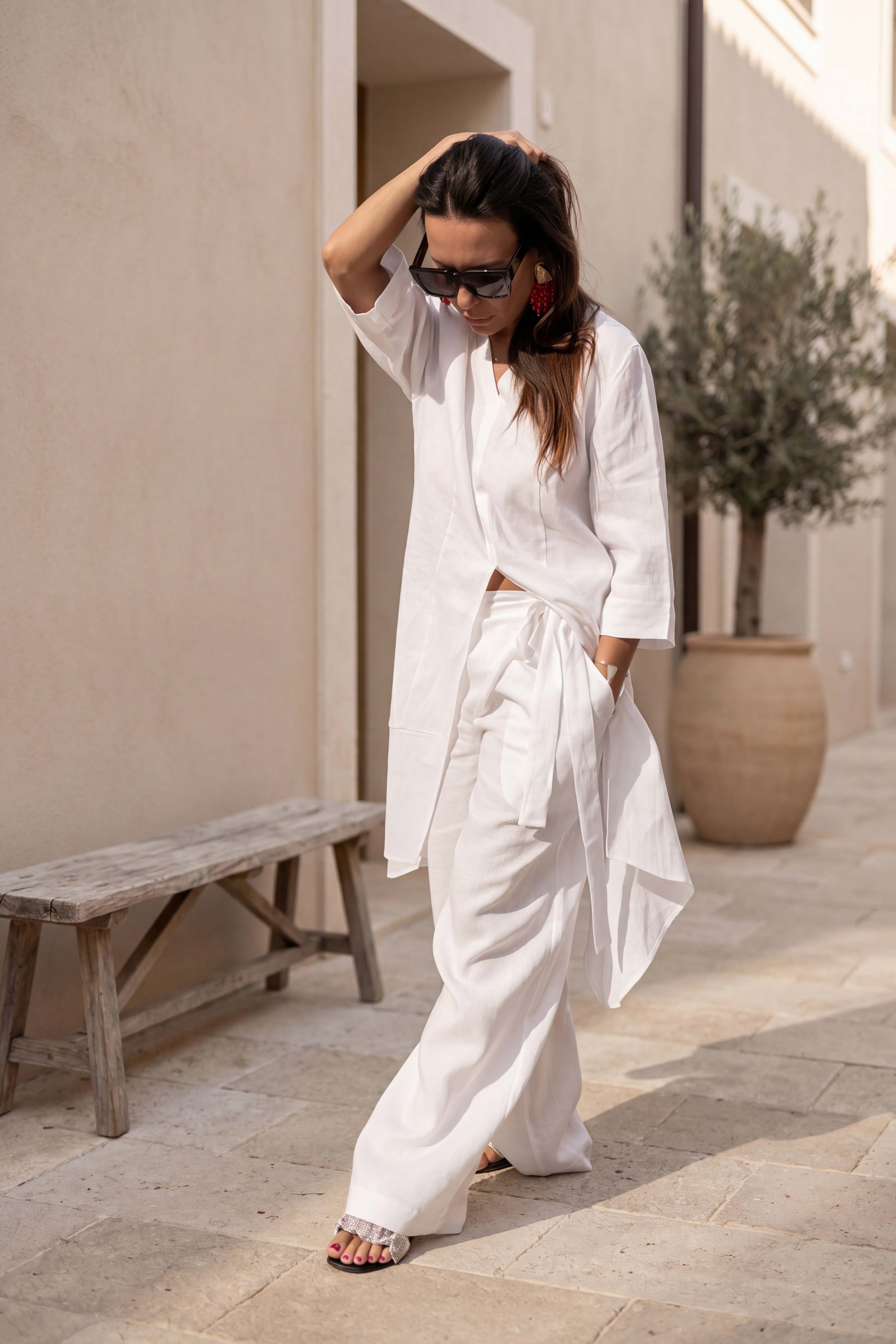 Woman in a white outfit standing outdoors near a building with a bench and plant.
