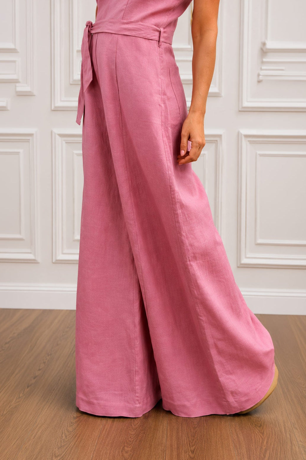 Person wearing a pink jumpsuit standing on a wooden floor with a white paneled wall in the background.