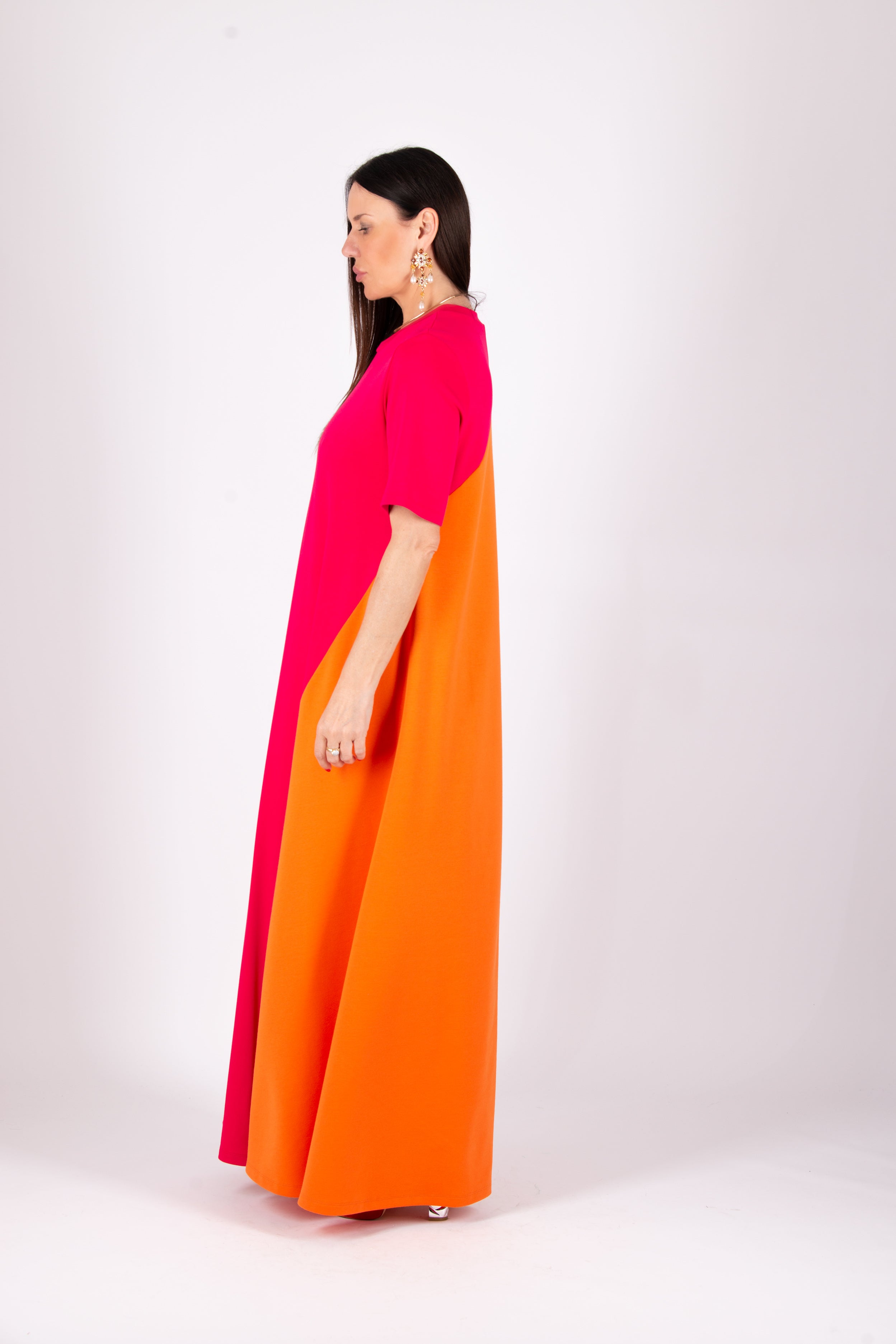 A woman modeling a long, color-blocked dress with a bright pink upper section and a vibrant orange lower skirt, shown from a side profile against a plain light gray studio background.