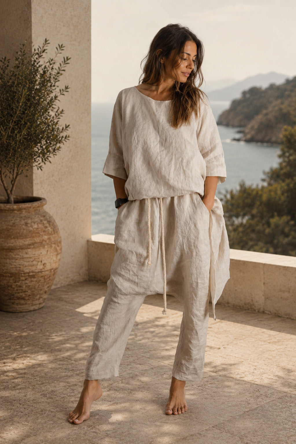 Woman in beige linen outfit standing on a balcony with a scenic view of water and mountains.