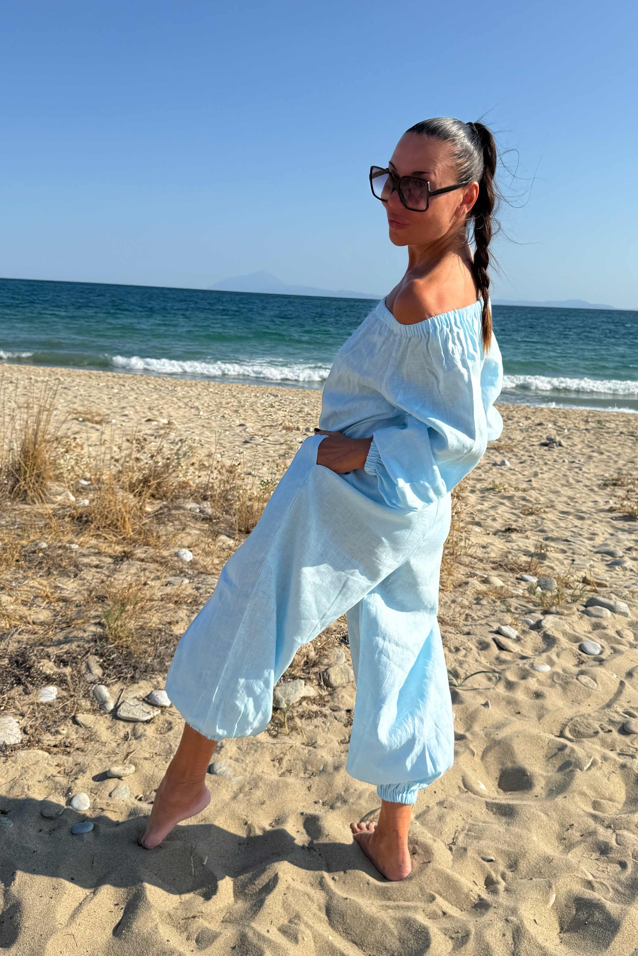 Woman in a light blue off-shoulder dress standing on a sandy beach with ocean in the background.