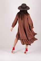 Woman wearing a brown dress with ruffles and a hat, standing against a white background