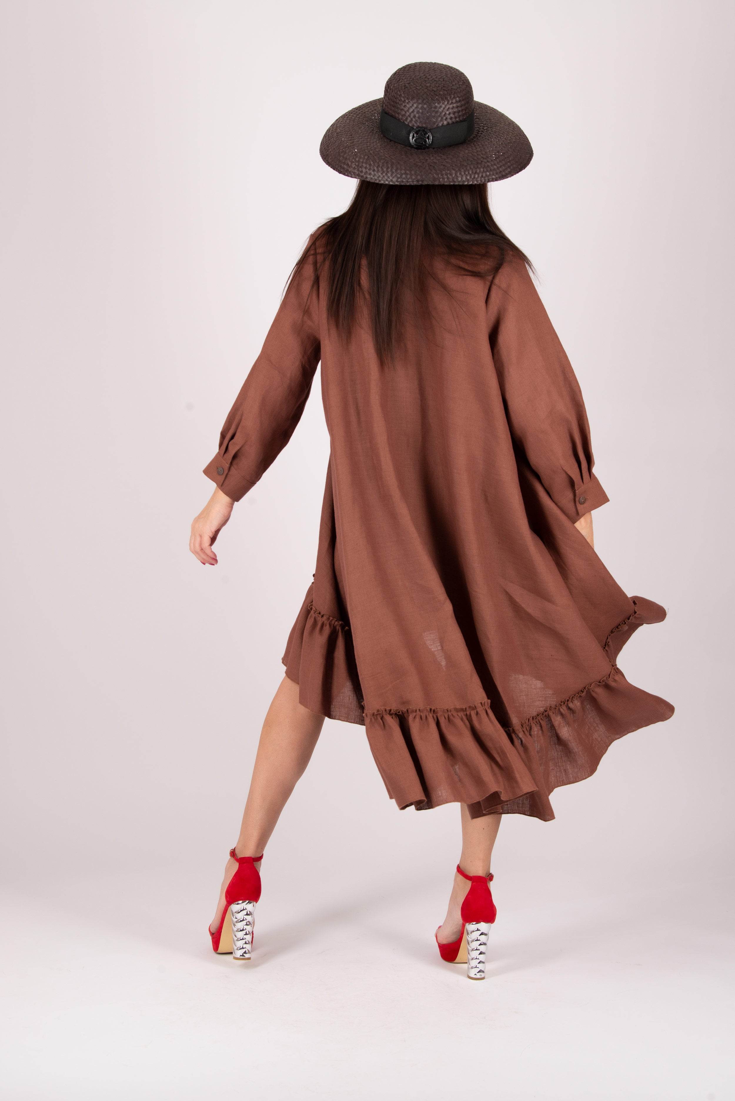 Woman wearing a brown dress with ruffles and a hat, standing against a white background
