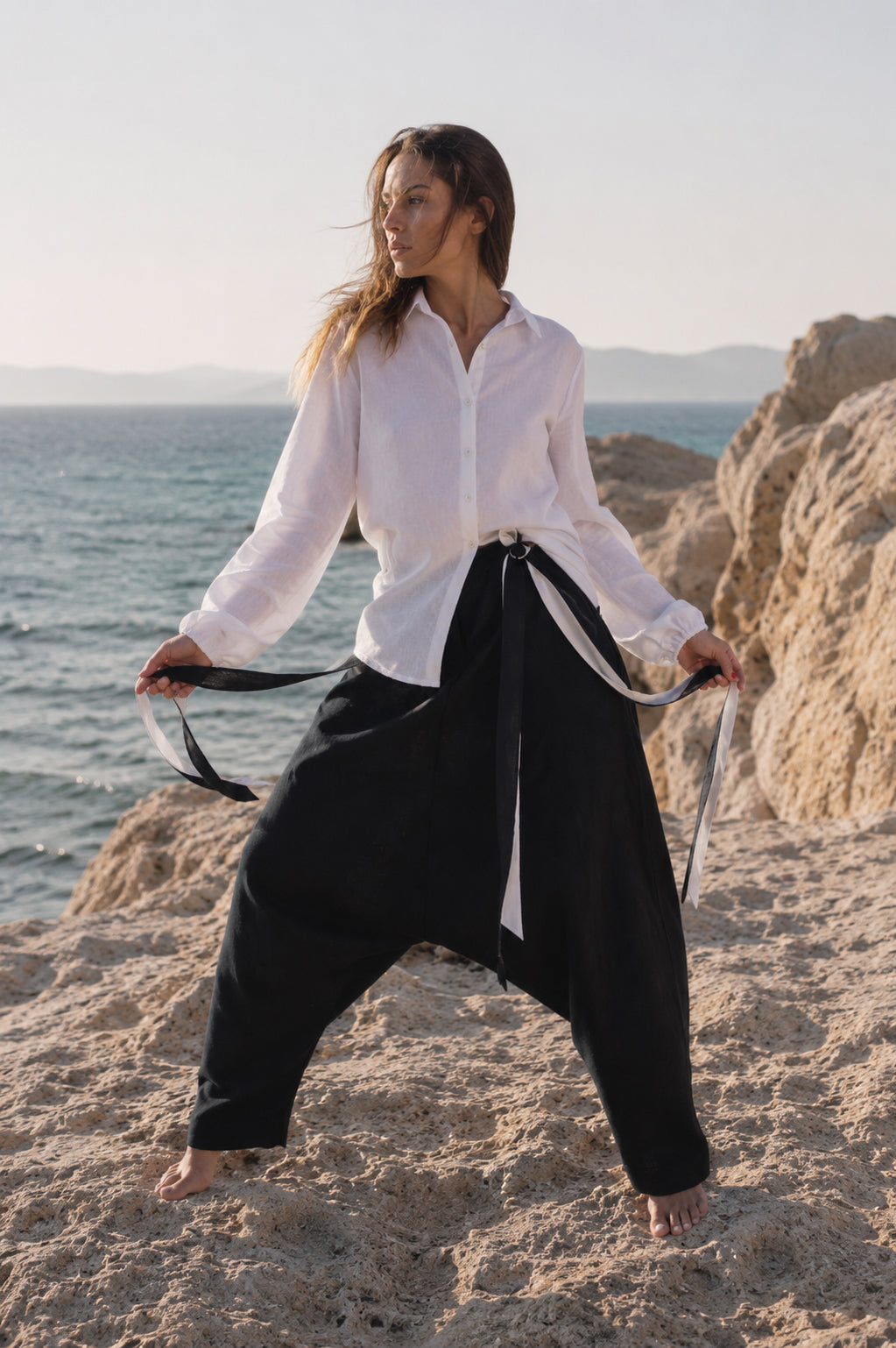 Woman in  black linen pants standing on a beach with ocean and rocks in the background