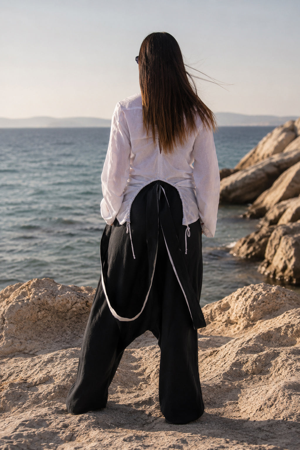 Person standing on rocks by the ocean wearing a white shirt and black pants.