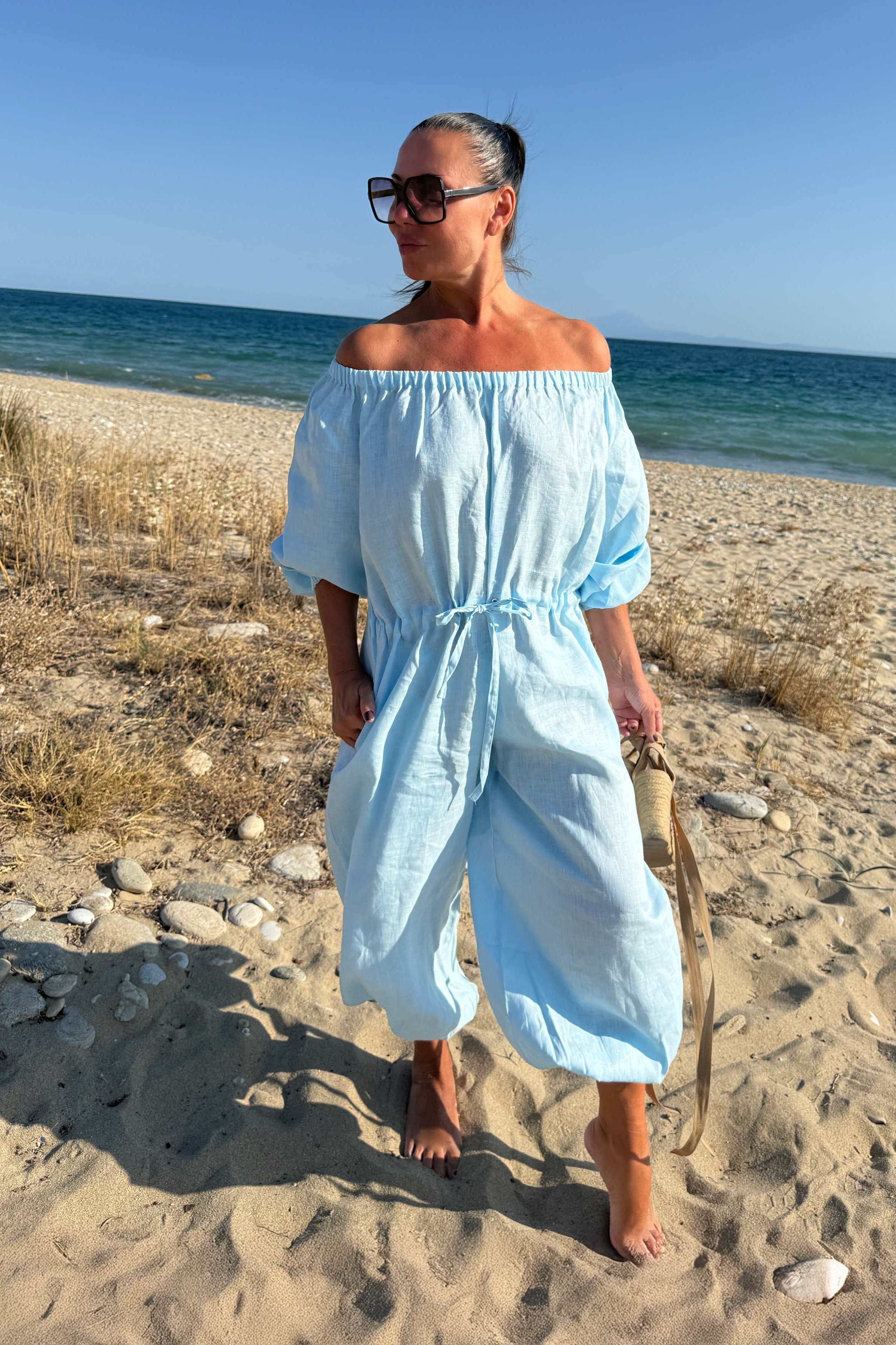 Woman in a light blue off-shoulder dress standing on a sandy beach with ocean in the background.
