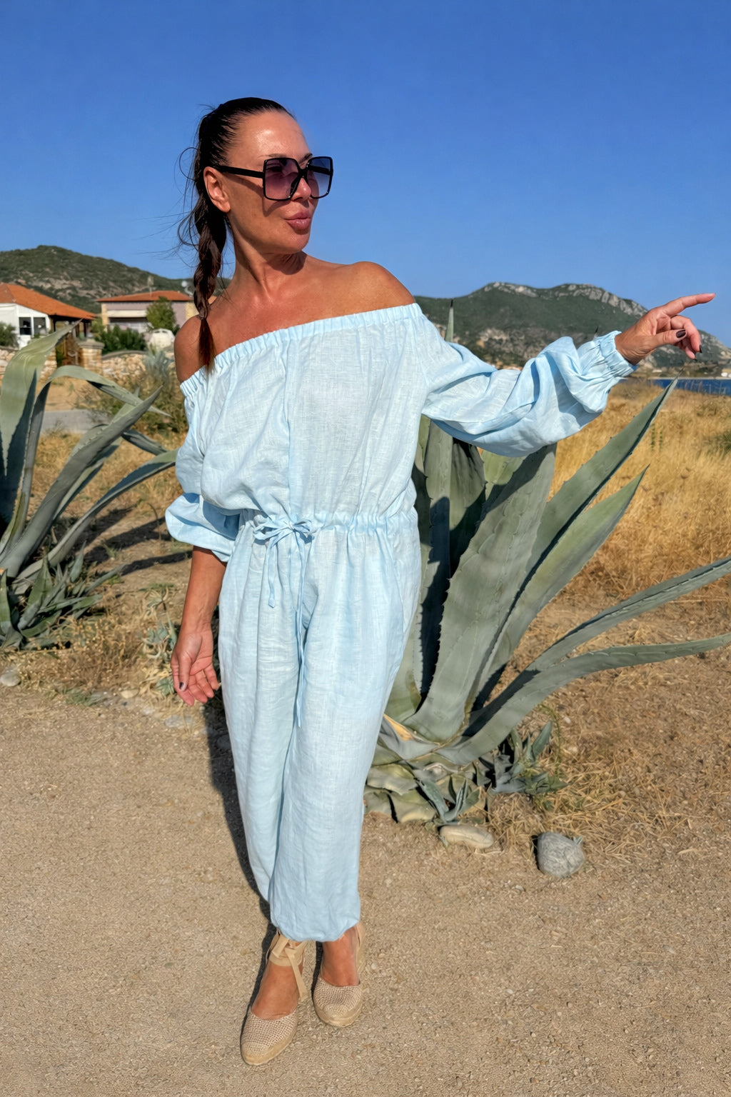 Woman in a light blue off-shoulder jumpsuit standing in a desert landscape with mountains in the background.
