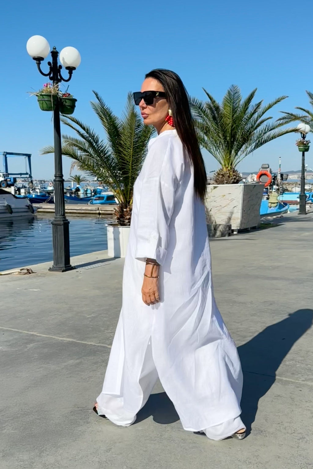 Woman in a white outfit standing by a waterfront with palm trees and a clear sky.