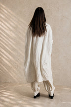 Person wearing a beige linen outfit standing against a beige wall with a soft shadow pattern.