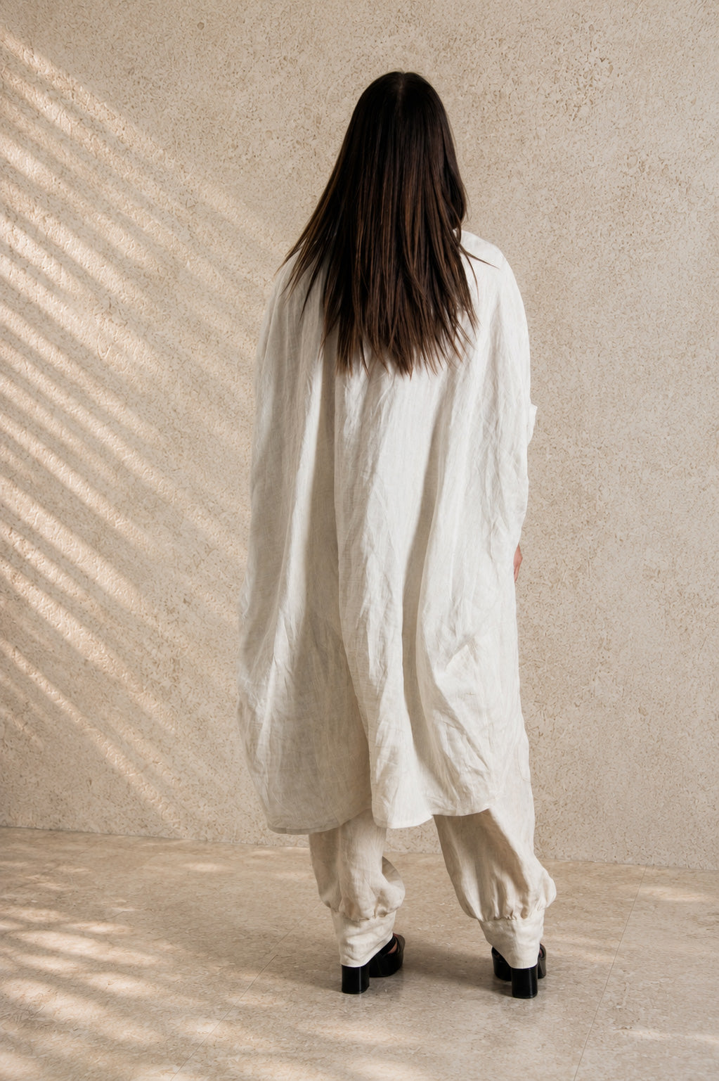 Person wearing a beige linen outfit standing against a beige wall with a soft shadow pattern.