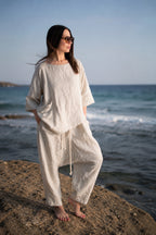 Woman in a loose-fitting beige outfit standing on a beach with ocean waves in the background