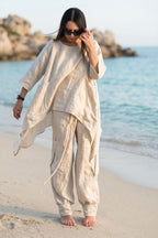 Woman in linen beige outfit standing on a beach with ocean and rocks in the background