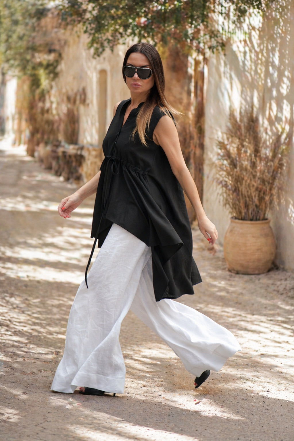 Woman in black sleeveless top and white pants walking on a sunlit path with potted plants.