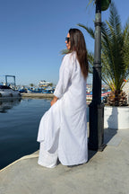 Woman in a white outfit standing by a waterfront with boats and palm trees in the background.