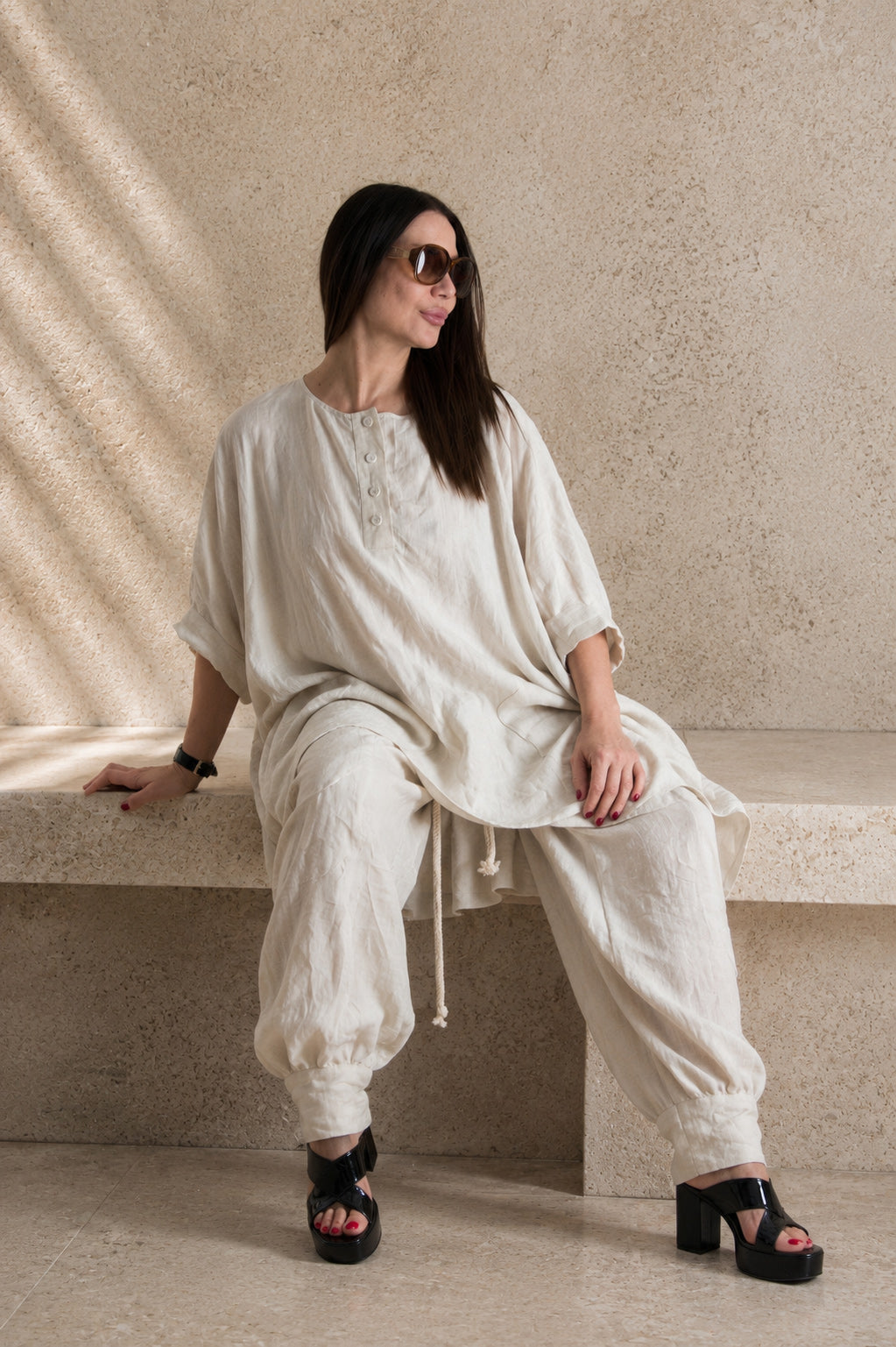 Woman in a natural beige linen outfit sitting on a stone bench with a neutral background