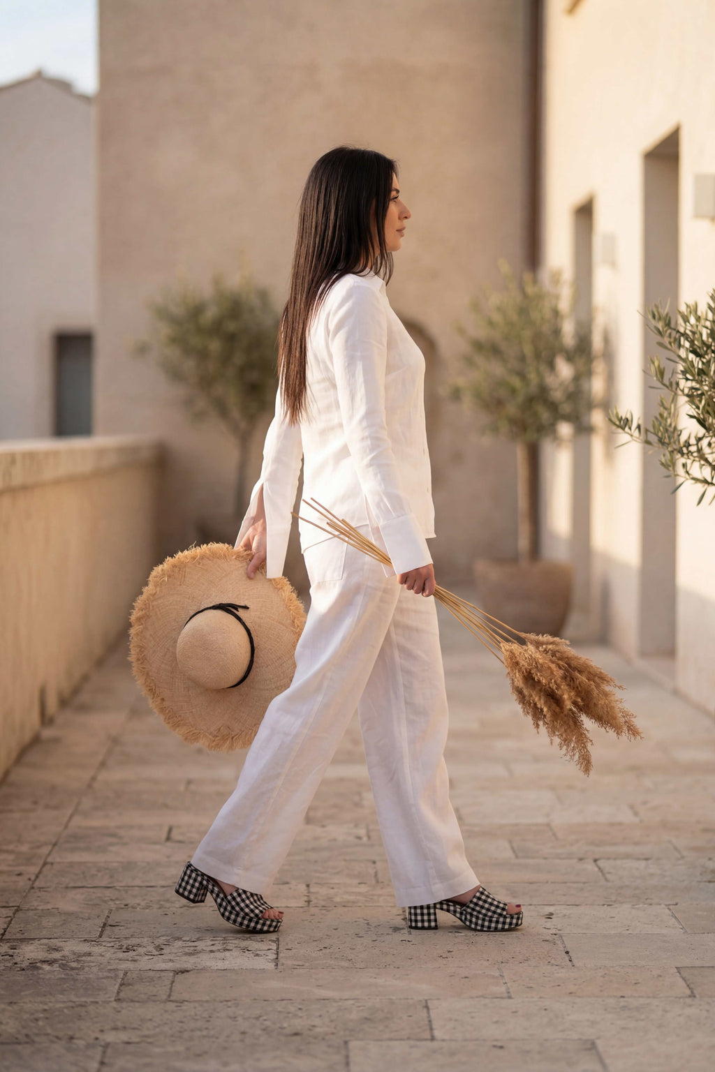 Woman in a white outfit walking outdoors with a straw hat and dried plants.
