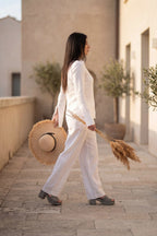 Woman in a white outfit walking outdoors with a straw hat and dried plants.