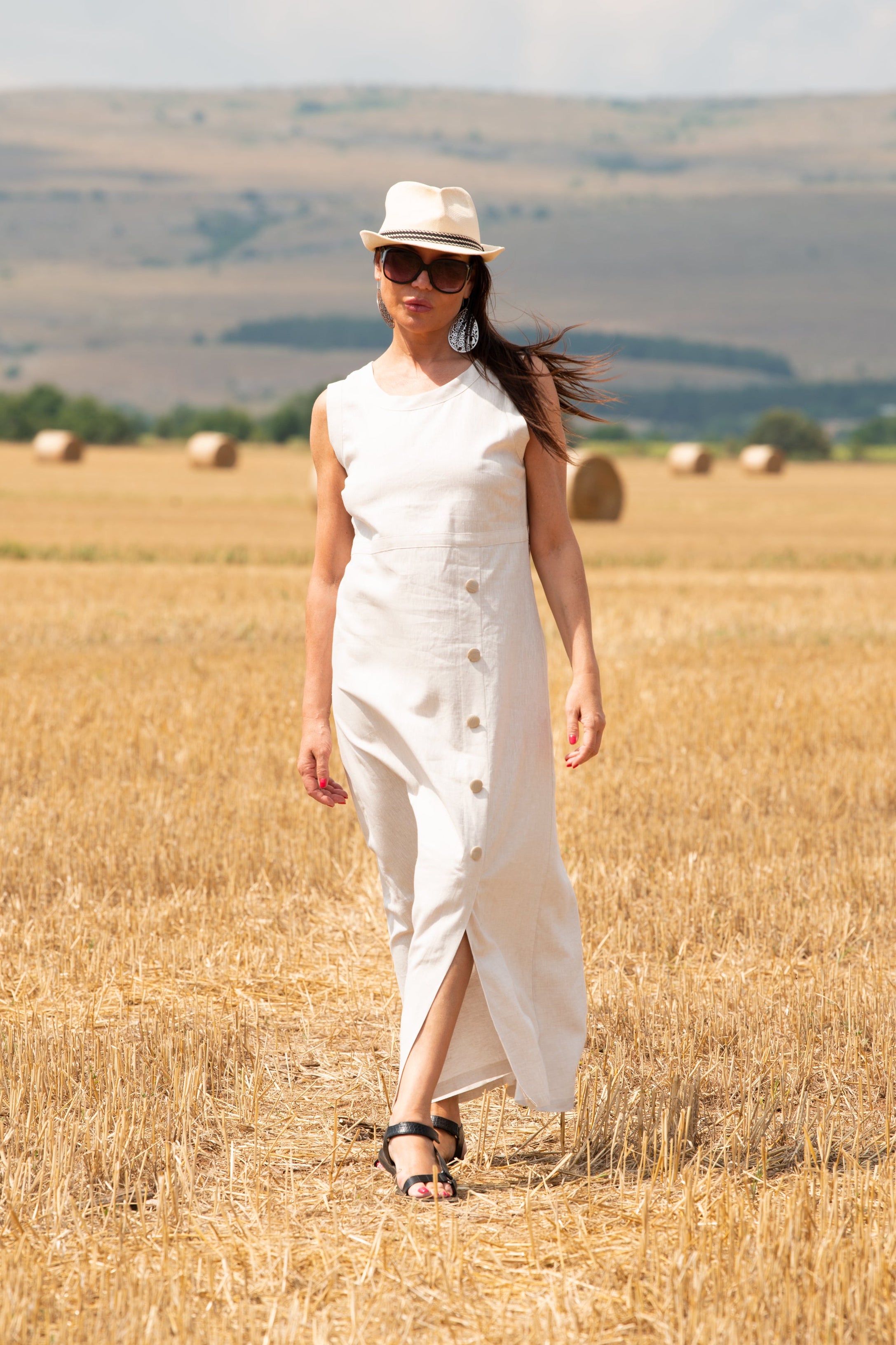 Woman in a white linen dress and hat standing in a field of straw bales.