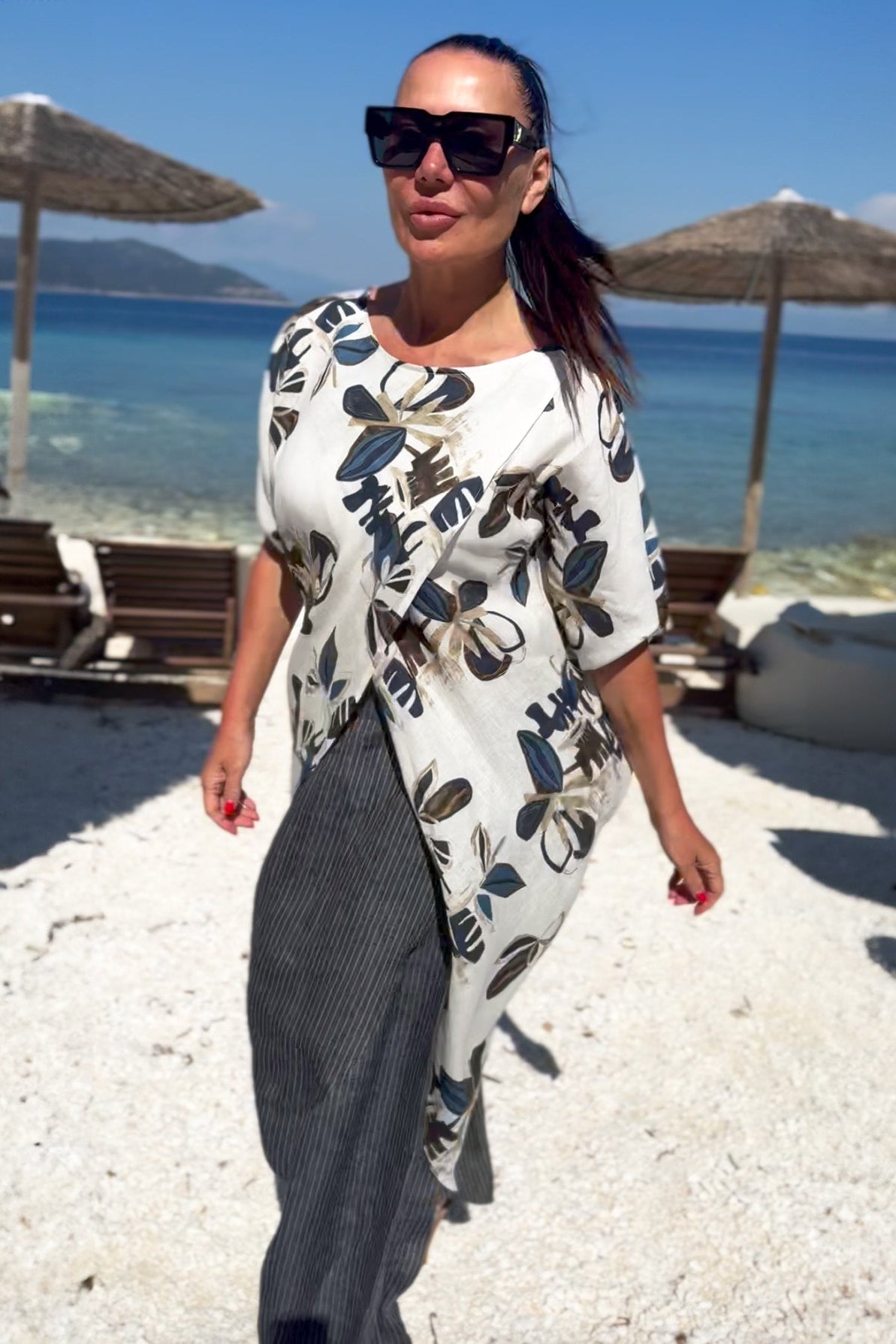 Woman in a floral tunic and sunglasses on a beach with umbrellas and chairs.